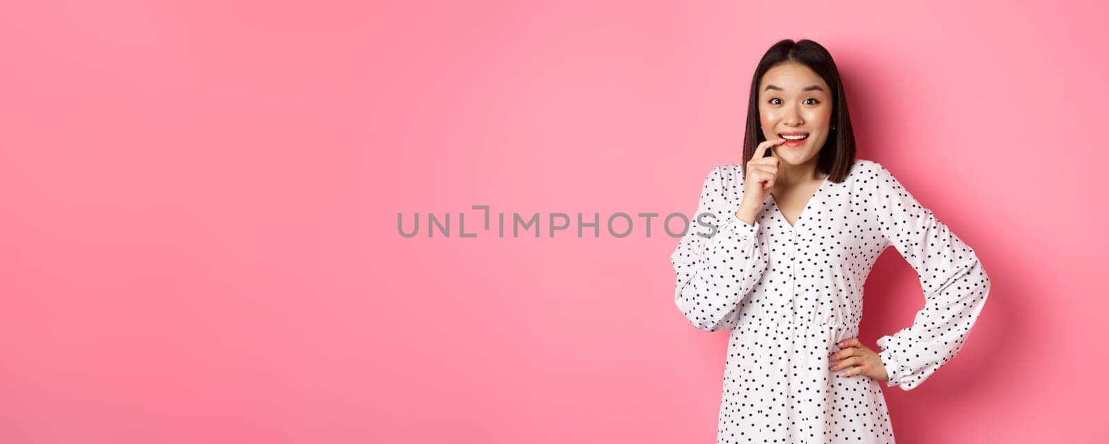 Image of beautiful young lady in dress, biting finger and smiling, staring with temptation and amazement, want buy something, standing over pink background.