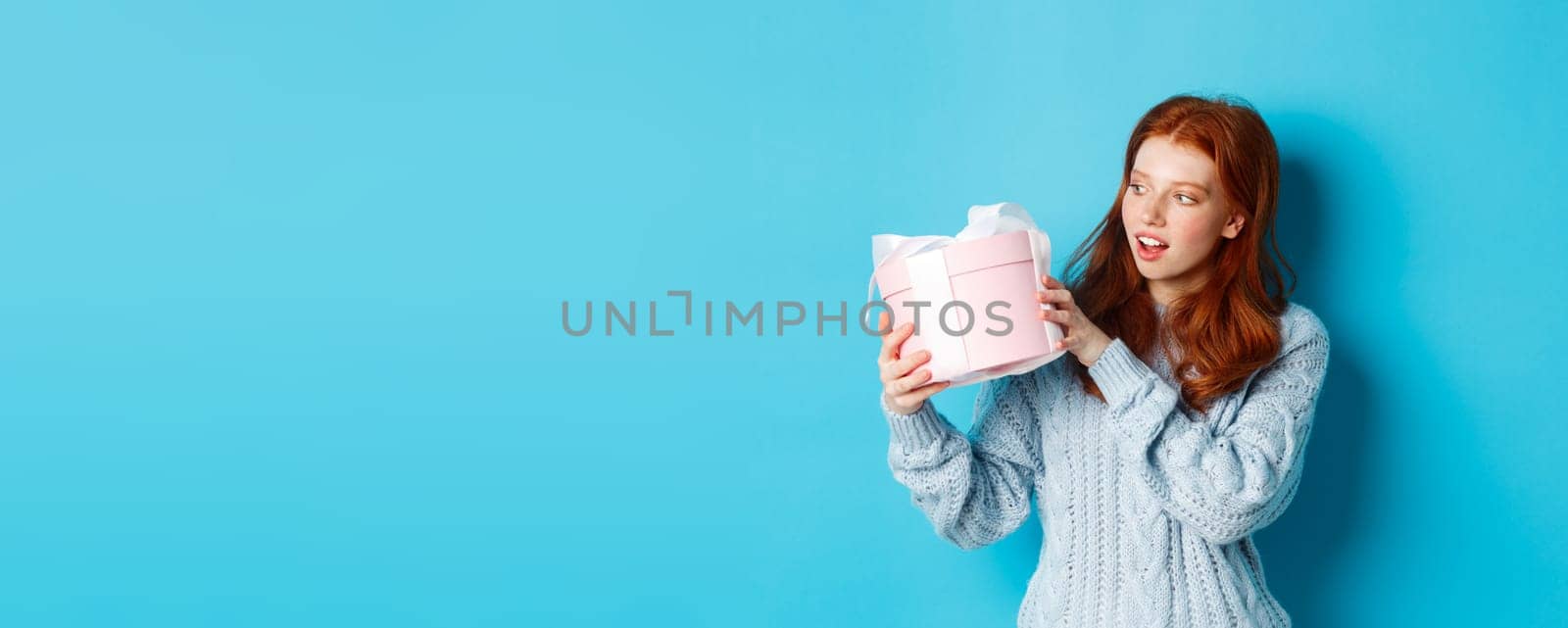 Curious teen girl with red hair, shaking gift box and wonder what inside, standing over blue background by Benzoix