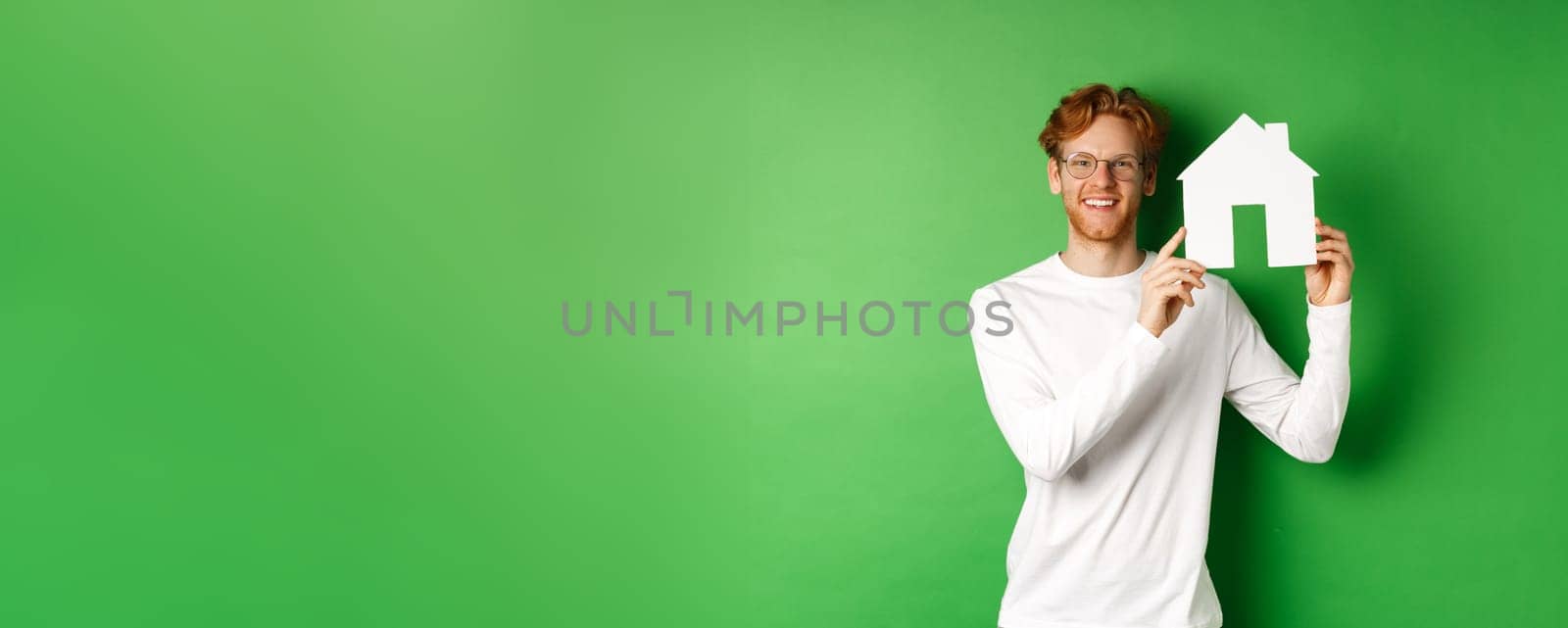 Real estate. Handsome young man with red hair, wearing glasses, showing paper house cutout and smiling, standing against green background by Benzoix