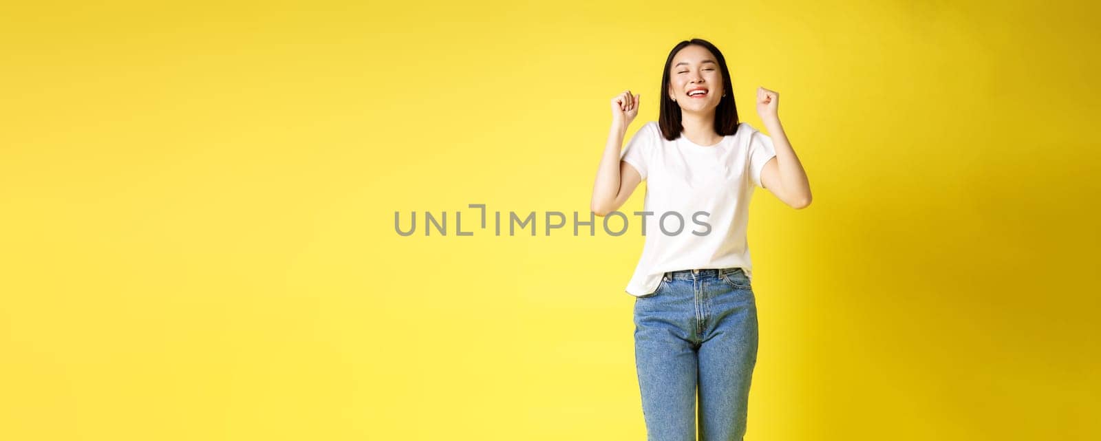 Full size shot of cheerful asian woman in jeans and white t-shirt jumping from happiness, winning prize and rejoicing, celebrating and shouting yes with joy, yellow background by Benzoix