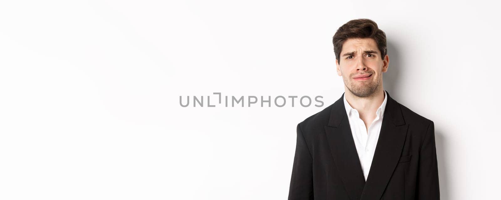 Close-up of confused handsome man in suit, grimacing and looking perplexed, wearing trendy suit, standing against white background.
