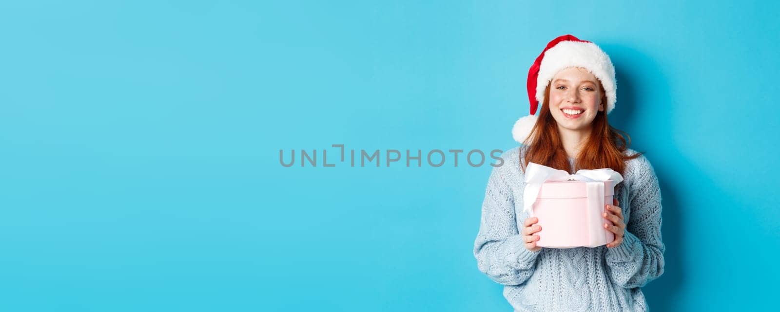 Winter holidays and Christmas Eve concept. Smiling redhead girl in sweater and Santa hat, holding New Year gift and looking at camera, standing against blue background.