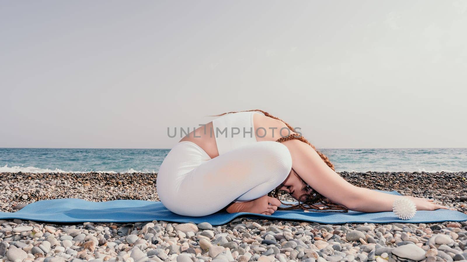 Woman yoga sea. Well looking middle aged woman with braids dreadlocks in white leggings and tops doing stretching pilates on yoga mat near sea. Female fitness yoga routine concept. Healthy lifestyle