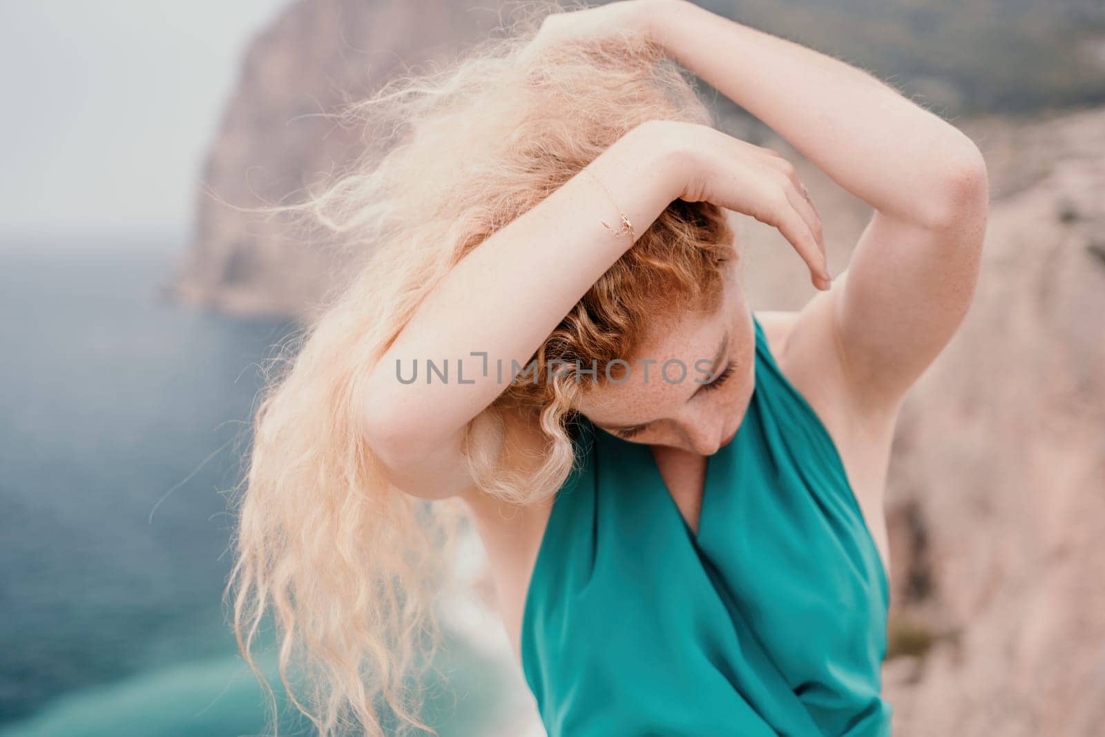 Redhead woman portrait. Curly redhead young caucasian woman with freckles looking at camera and smiling. Close up portrait cute woman in a mint long dress posing on a volcanic rock high above the sea by panophotograph