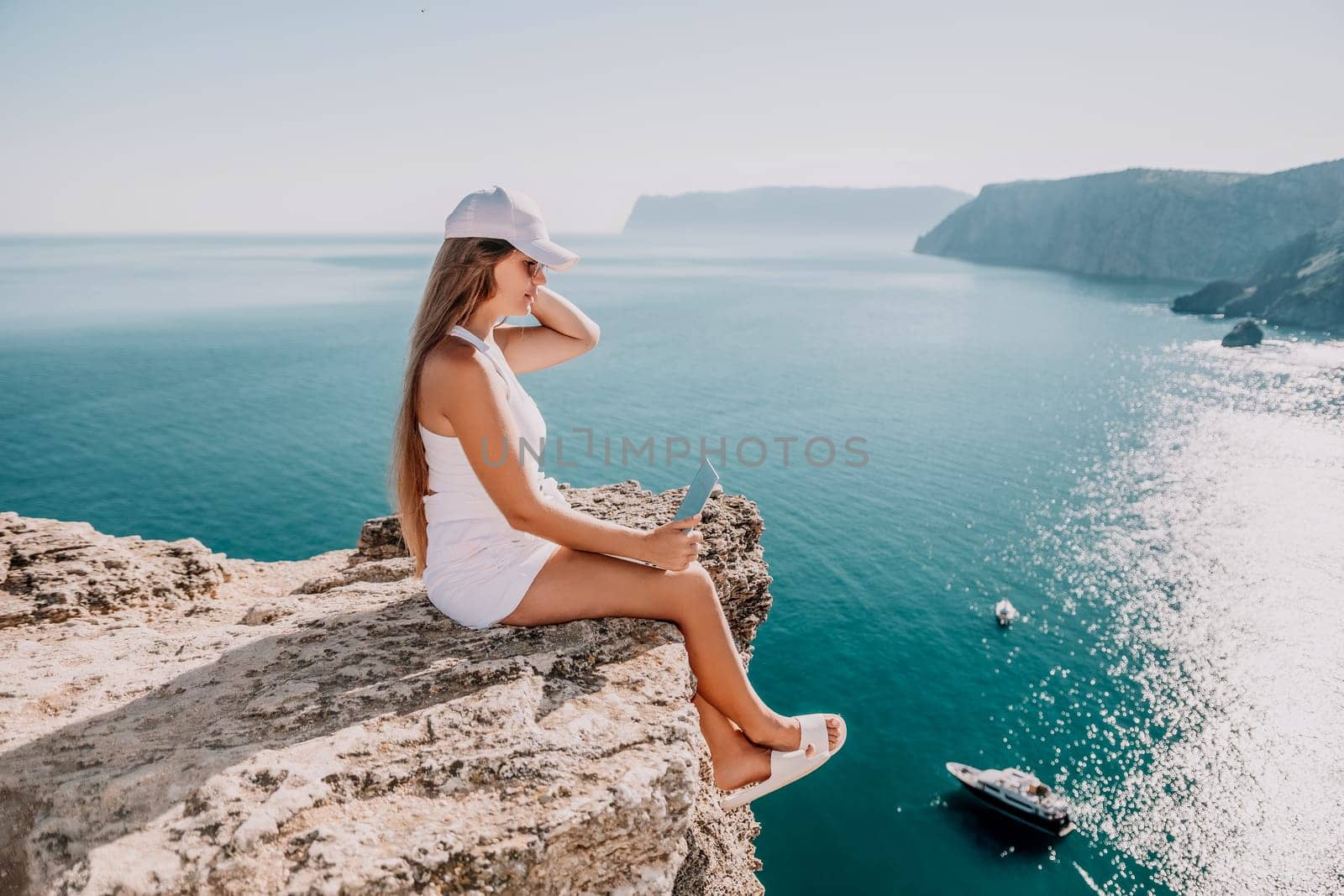 Successful business woman in yellow hat working on laptop by the sea. Pretty lady typing on computer at summer day outdoors. Freelance, travel and holidays concept.