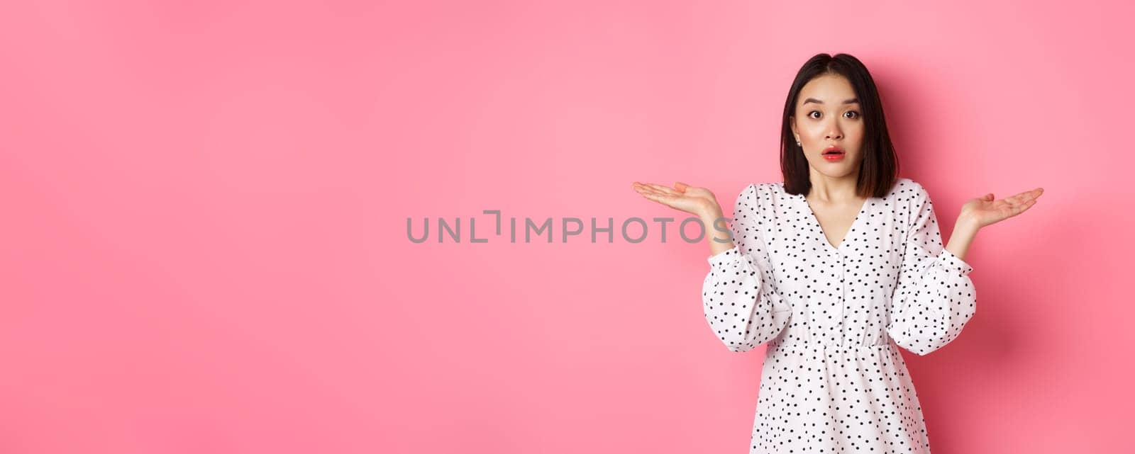 Confused and clueless asian girl shrugging, staring at camera puzzled, dont know what to do, spread hands sideways, standing over pink background by Benzoix