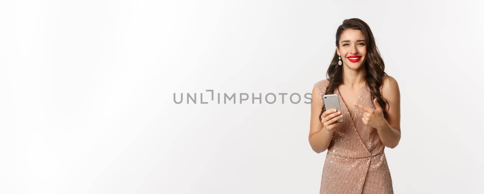 Holidays, online shopping concept. Satisfied good-looking woman in luxury dress and red lipstick, pointing at mobile phone, smiling pleased, white background.