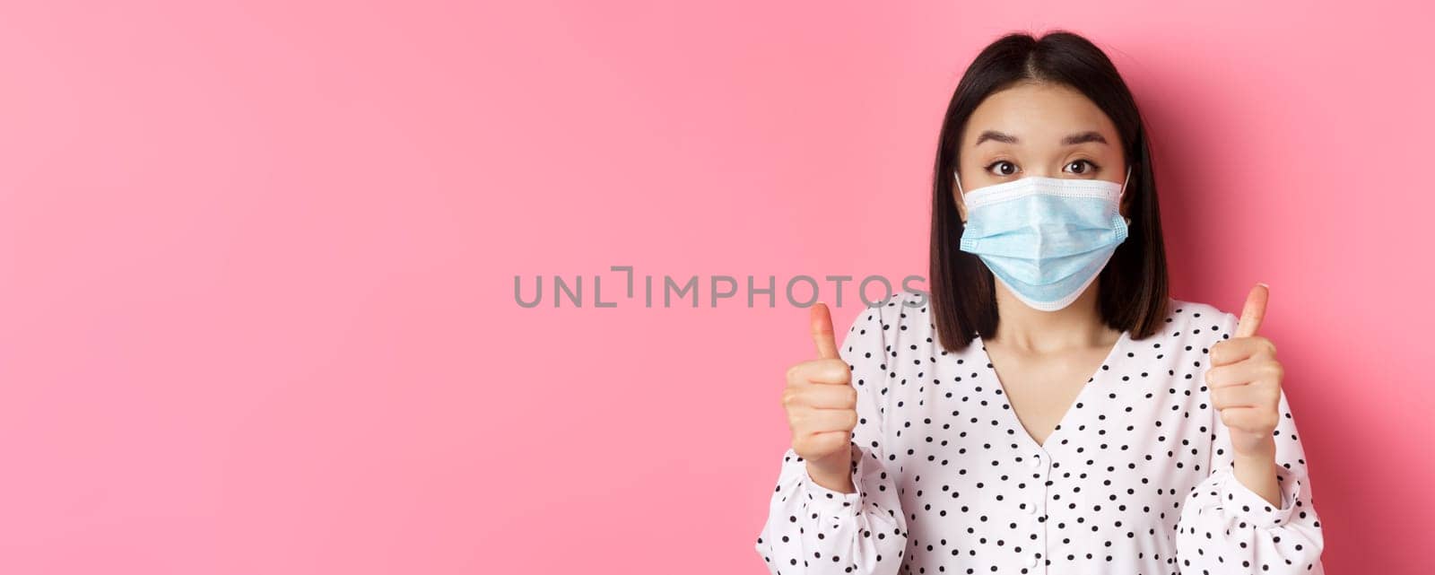 Covid-19, pandemic and lifestyle concept. Supportive asian female in face mask showing thumbs-up, approve and praise good work, standing over pink background.
