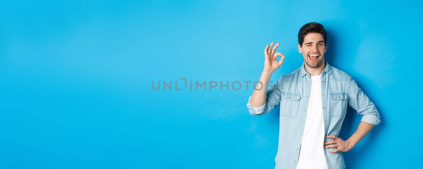 Confident adult man winking and showing okay sign, approve something good, standing against blue background by Benzoix