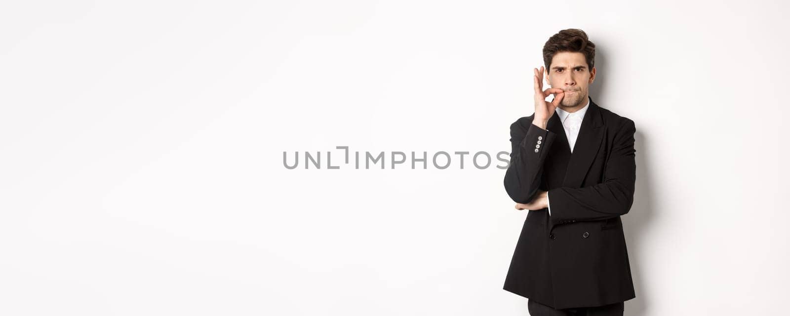 Image of serious businessman in black suit, zipping mouth and frowning, hiding secret, showing taboo sign, standing over white background.