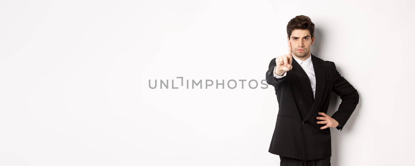Portrait of serious handsome man in business suit, showing one finger to prohibit or decline something, telling to stop, disagree with you, standing over white background.
