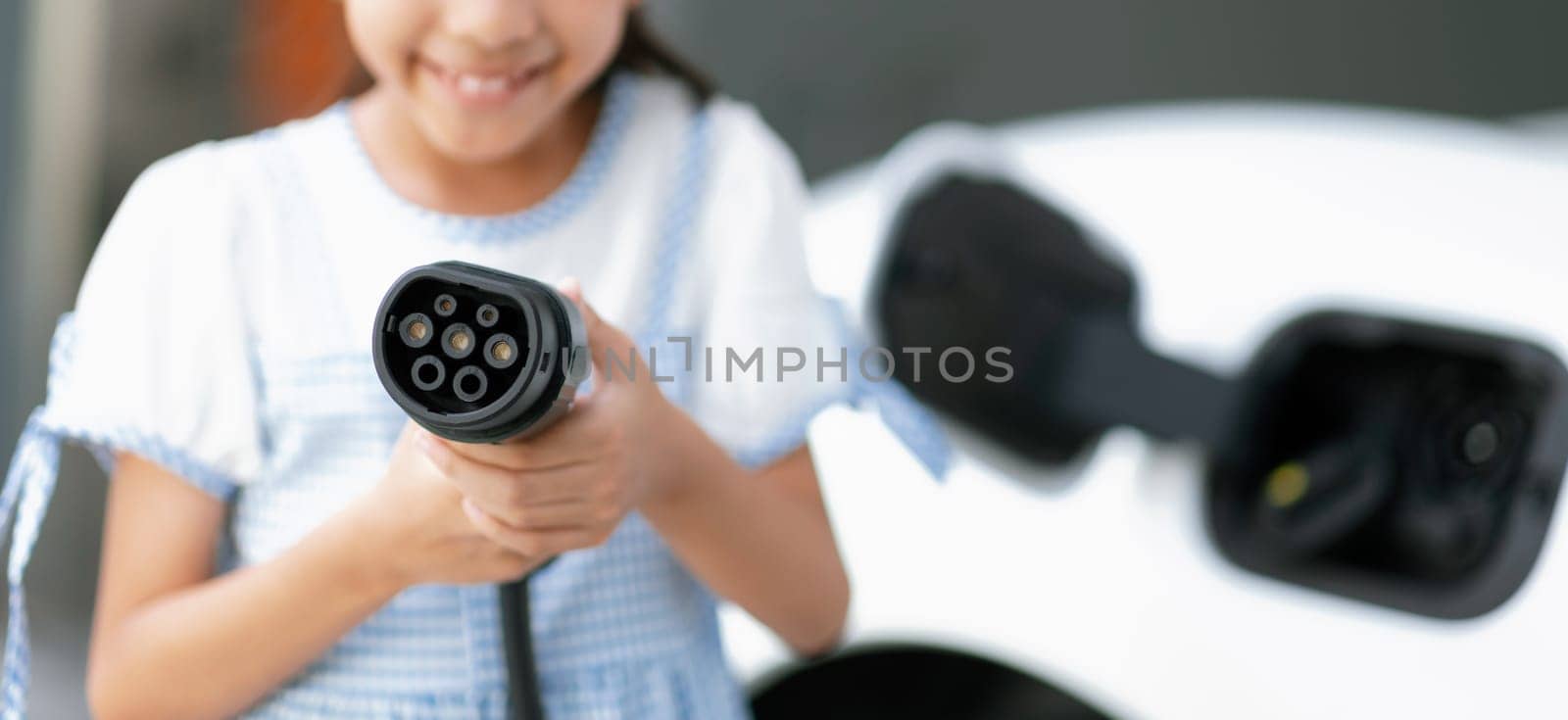 Focus electric charger plug from home charging station for EV engine power by renewable energy with blur background of girl pointing charger device at camera. Concept of progressive young generation.