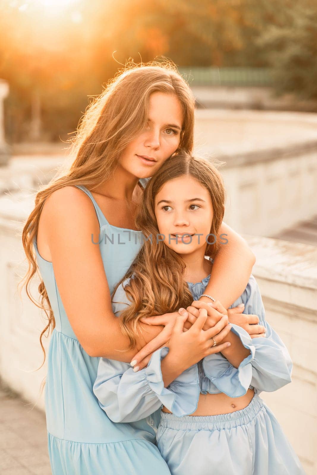 Portrait of mother and daughter in blue dresses with flowing long hair against the backdrop of sunset. The woman hugs and presses the girl to her. They are looking at the camera. by Matiunina