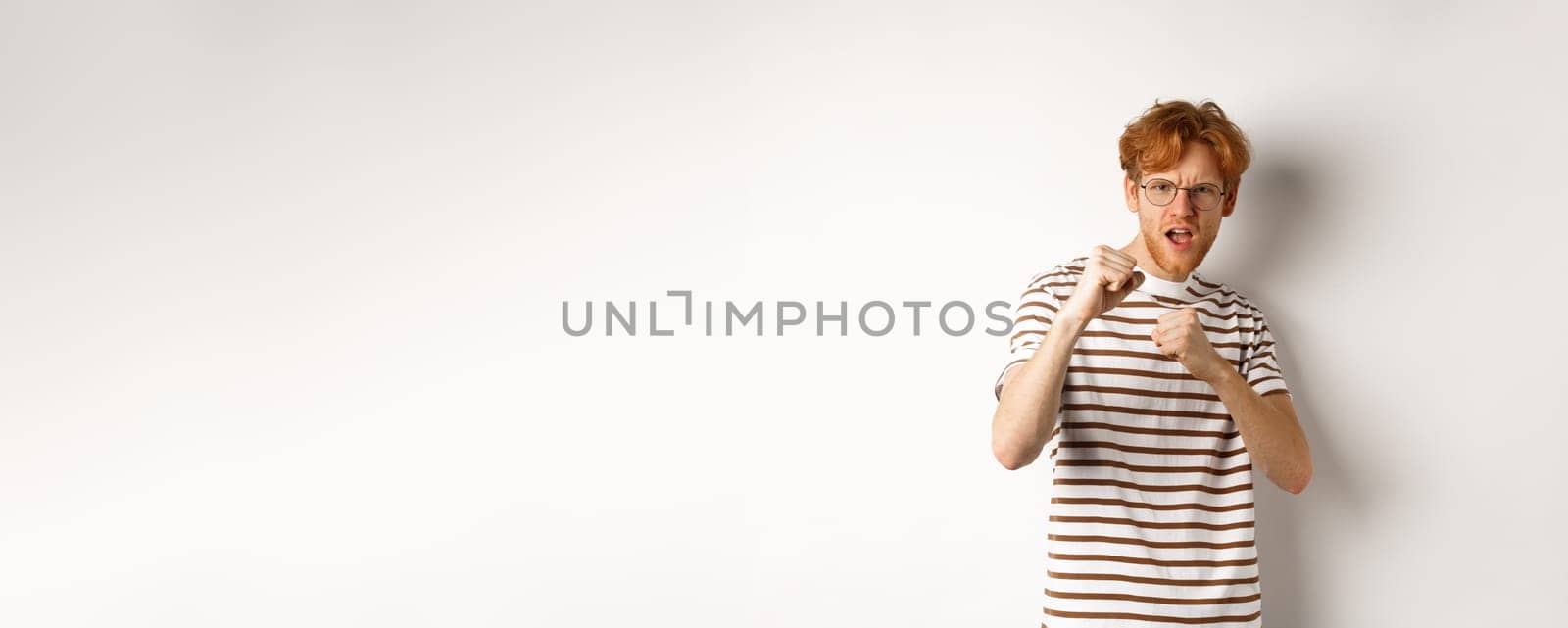 Funny young man with red hair raising fists for fight, shadow boxing and looking serious at camera, standing over white background.