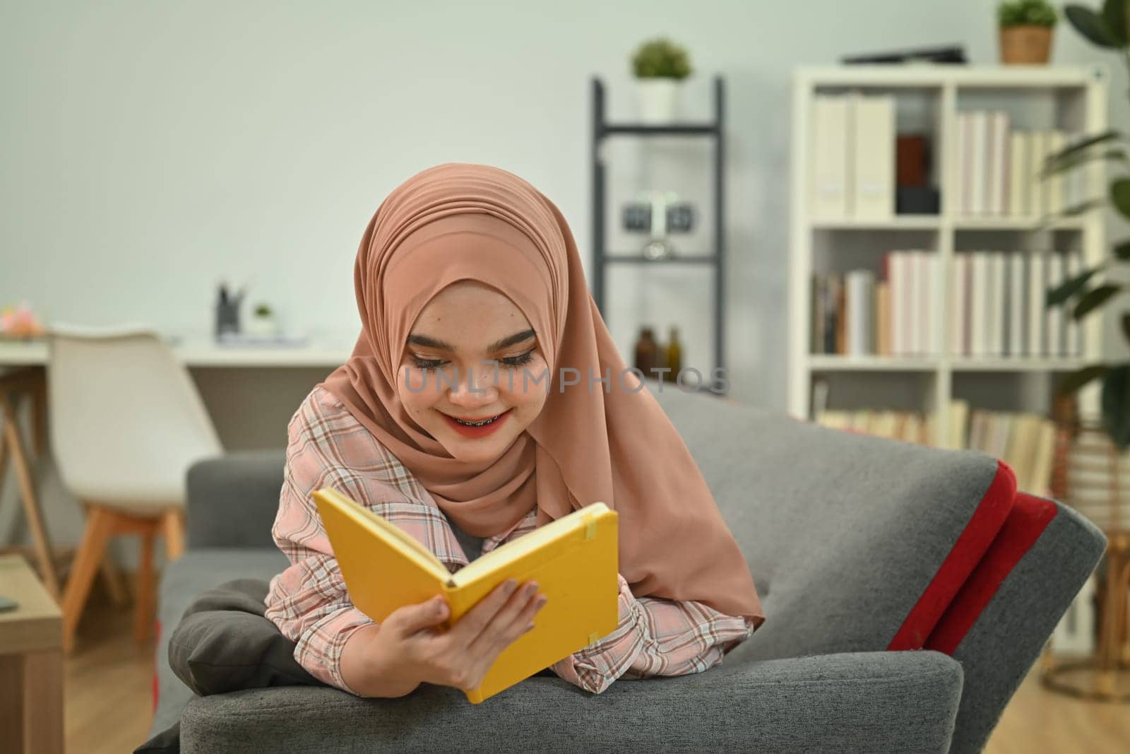 Pleasant millennial Muslim woman in hijab reading book, laying on couch in living room. People and leisure activity concept.