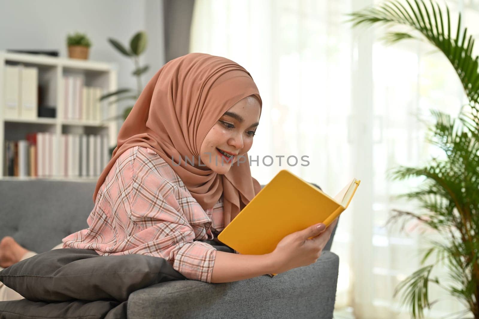 Happy young Muslim woman in hijab and casual clothes laying on couch and reading a book. People and leisure activity concept.