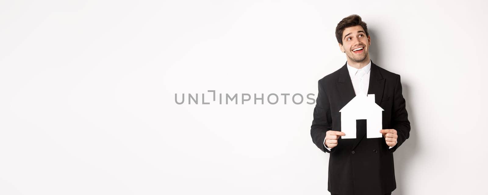 Portrait of handsome man in suit searching for home, holding paper house and looking at upper right corner dreamy, standing over white background.