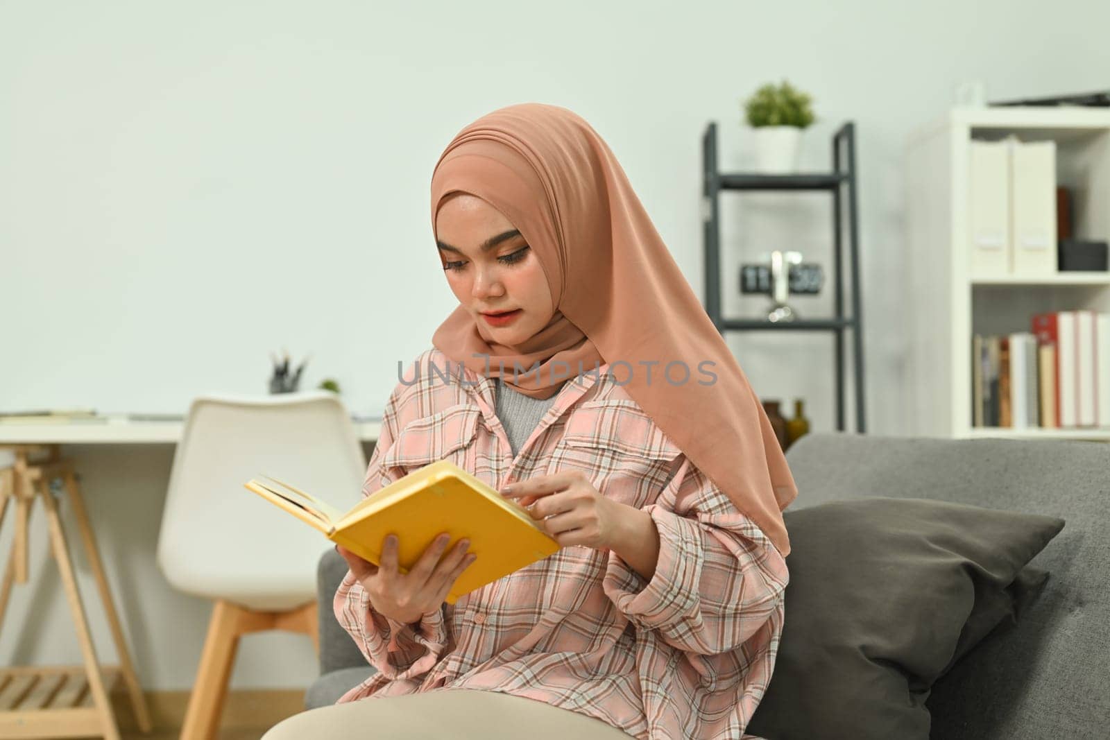 Calm young muslim woman wearing hijab reading book on couch, spending leisure time at home.