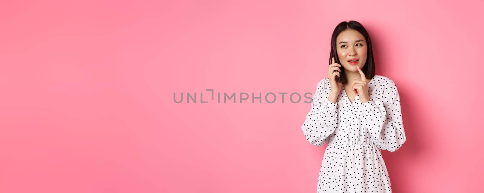 Thoughtful asian woman calling on mobile phone to make order, thinking and looking aside, standing against pink background by Benzoix