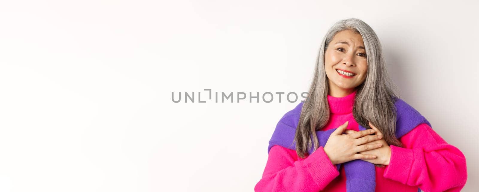 Close up of stylish asian mother holding hands on heart, looking touched and heartfelth, saying thank you, smiling grateful, standing over white background by Benzoix