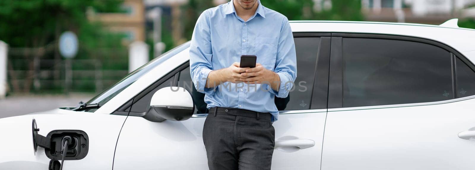 Progressive businessman talking on the phone, leaning on electric car recharging with public EV charging station, apartment condo residential building on the background as green city lifestyle.