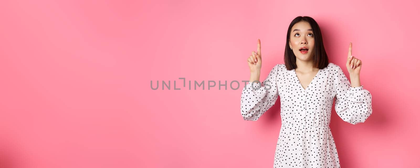 Image of beautiful asian girl in dress looking, pointing fingers up, gazing dreamy at promo offer, standing over pink background by Benzoix