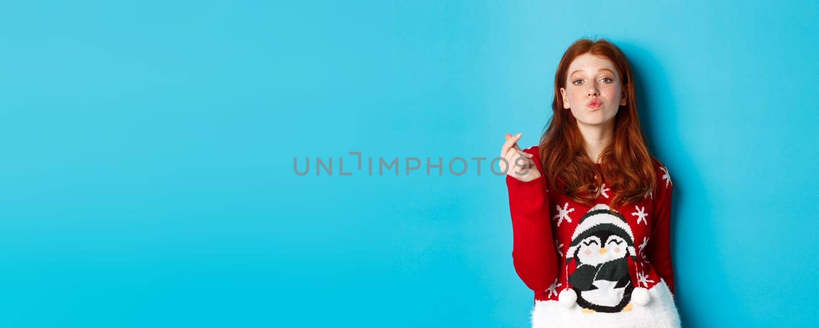 Winter holidays and Christmas Eve concept. Lovely redhead girl in xmas sweater, showing heart sign and pucker lips for kiss, standing over blue background by Benzoix
