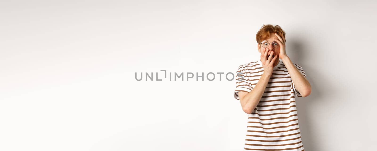 Scared redhead guy screaming terrified, holding hands on face from fear, standing over white background.