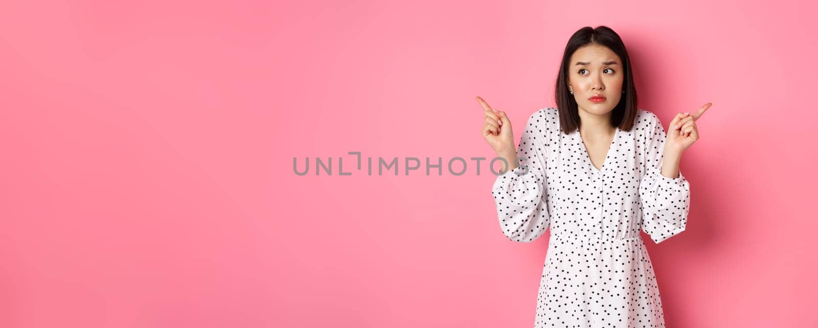 Confused asian woman hesitating, shrugging and pointing hands two sides, staring indecisive and need help with choice, standing over pink background by Benzoix