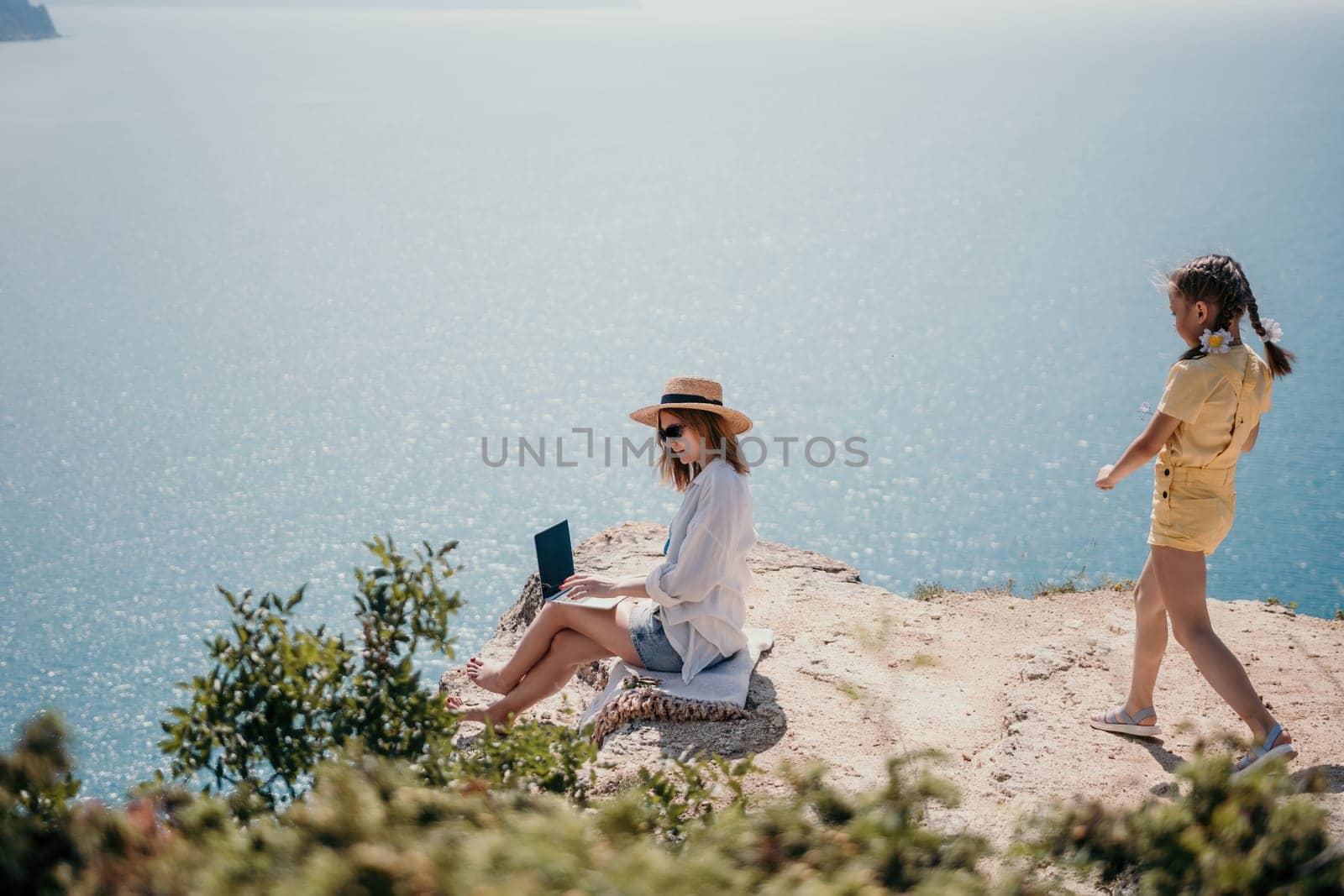 Successful business woman in yellow hat working on laptop by the sea. Pretty lady typing on computer at summer day outdoors. Freelance, travel and holidays concept.