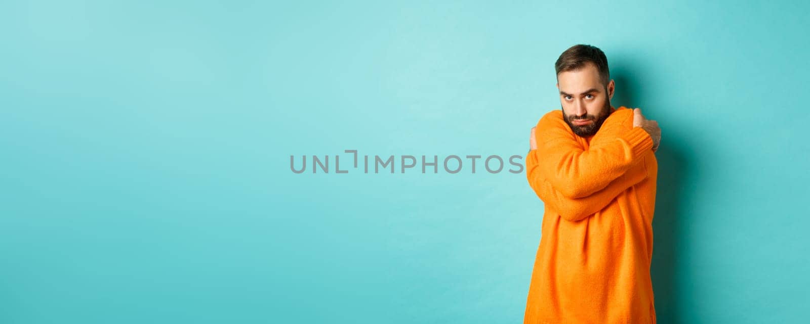 Timid man feeling offended and defensive, hugging himself and looking suspicious at camera, standing over light blue background.