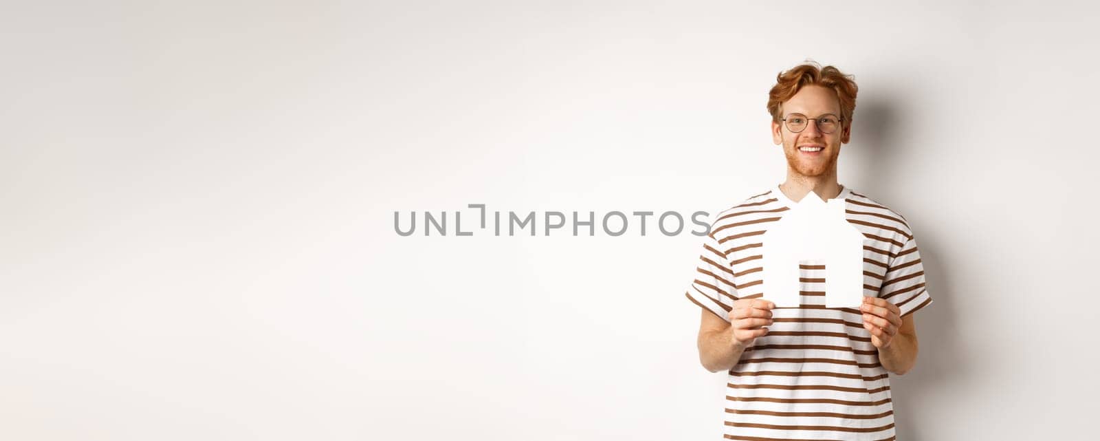 Real estate concept. Smiling redhead man in glasses holding cutout paper house and looking at camera, saving for buying property, standing over white background.