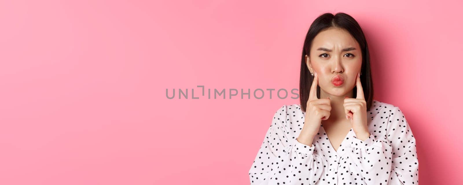 Beauty and lifestyle concept. Cute asian woman making grumpy face, poking cheeks and grimacing, standing over pink background.