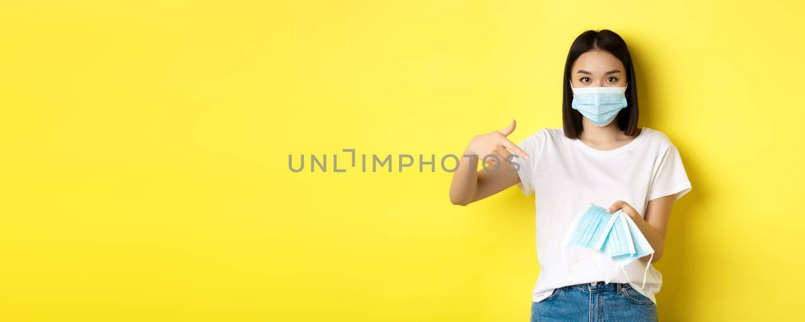 Coronavirus, quarantine and medicine concept. Young asian woman pointing finger at medical masks for going outdoors, standing over yellow background by Benzoix
