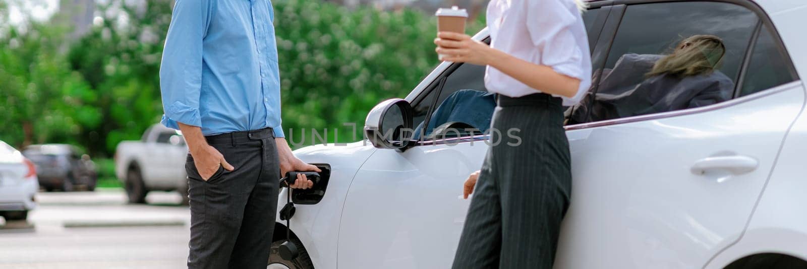 Progressive businessman and businesswoman with coffee, standing at electric car connected to charging station before driving around city center. Eco friendly rechargeable car powered by clean energy.