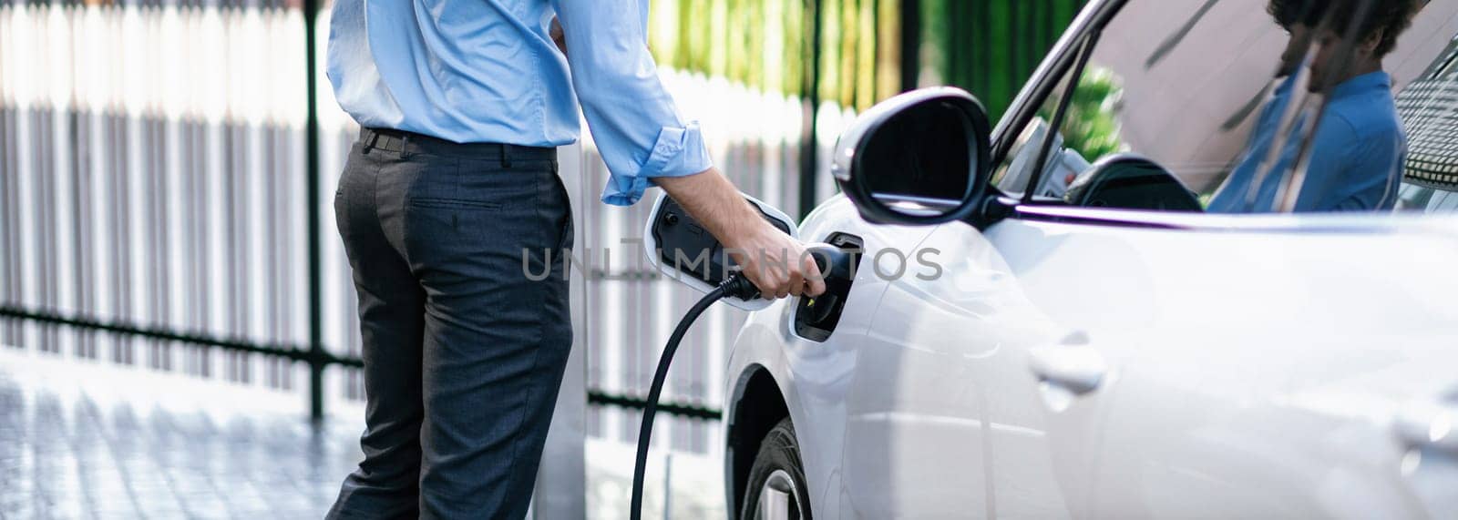 Closeup progressive man holding EV charger plug from public charging station for electric vehicle with background of residential building as concept eco-friendly sustainability energy car concept.