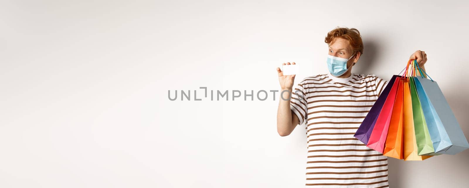 Pandemic and special offer concept. Happy redhead man in face mask showing shopping bags and plastic credit card, standing over white background by Benzoix