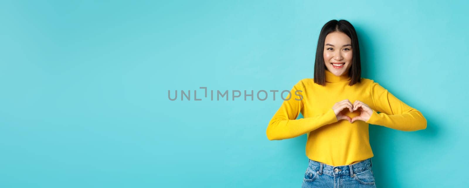 Valentines day and romance concept. Beautiful asian woman show I love you, heart gesture and smiling, standing against blue background.