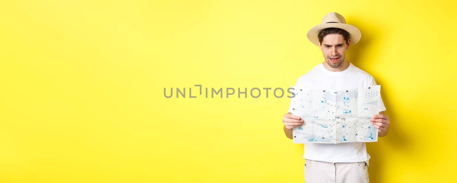 Travelling, vacation and tourism concept. Man looking confused at map during trip, cant understand, standing over yellow background by Benzoix