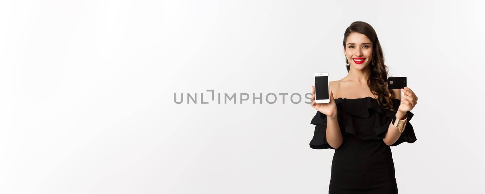 Online shopping concept. Fashionable brunette woman in black dress, showing mobile screen and credit card, smiling pleased, standing over white background by Benzoix