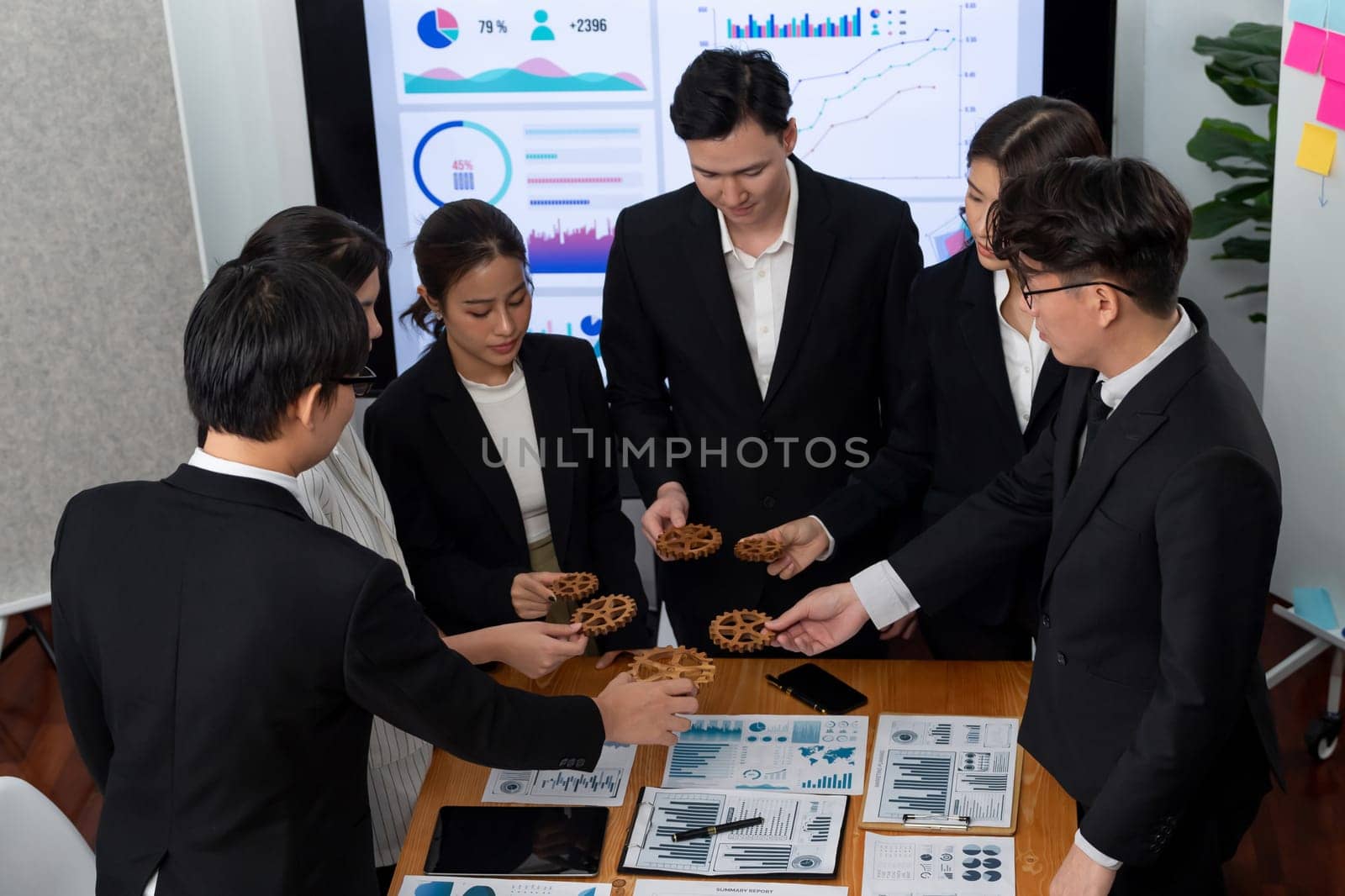 Businesspeople wearing suit for harmony synergy in office workplace concept hand holding gears into collective form with data analyzed by Bi in dashboard report papers on table.