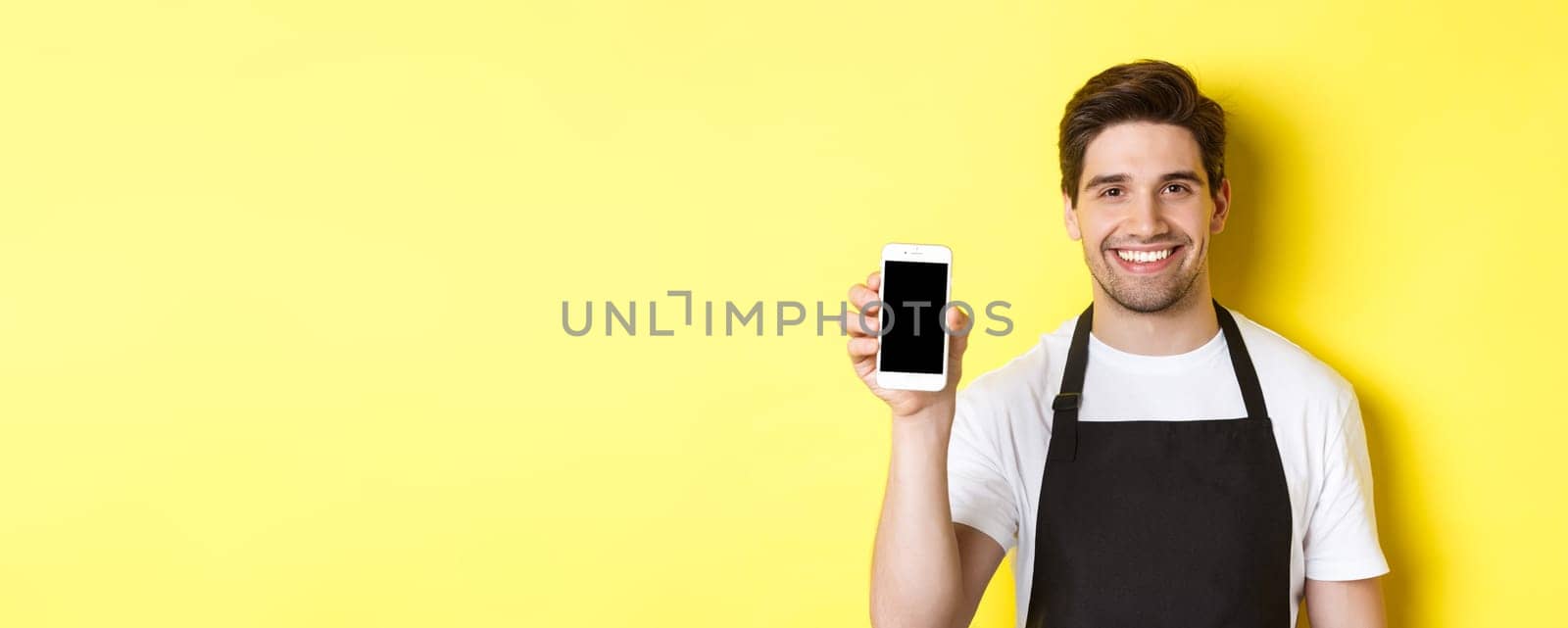Close-up of handsome waiter in black apron showing smartphone screen, recommending app, standing over yellow background by Benzoix