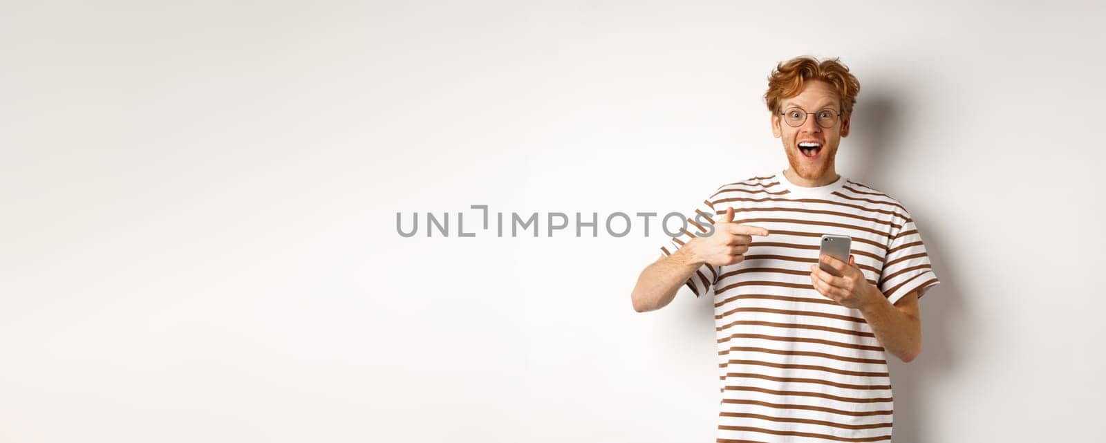 Technology and e-commerce concept. Amazed redhead man checking out online promo offer on mobile phone, pointing finger at smartphone and smiling, white background by Benzoix