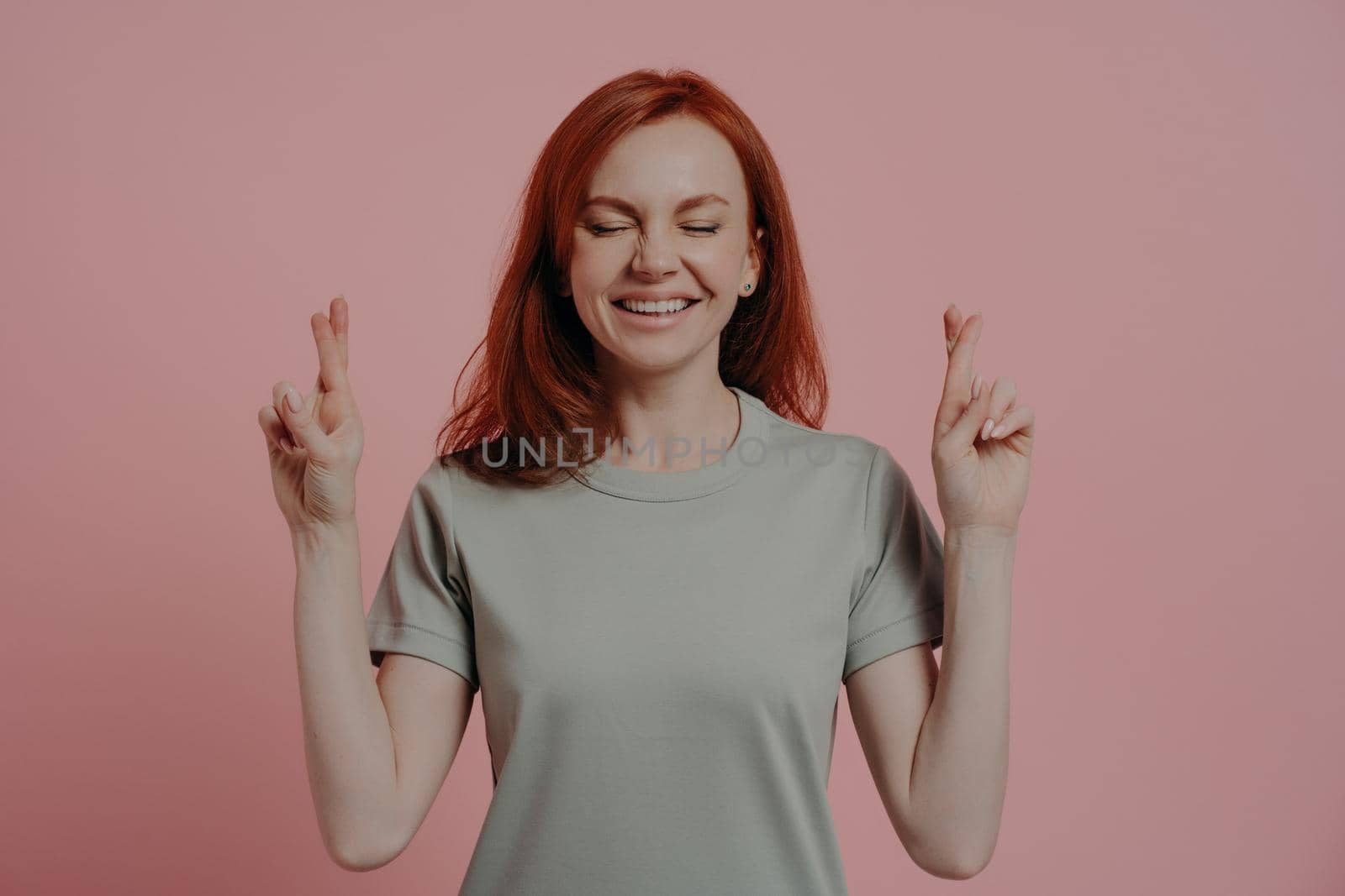 Studio shot of hopeful red-haired female student with closed eyes in casual wear crossing fingers with superstitious facial expression, asking for good luck before exam, isolated over pink background