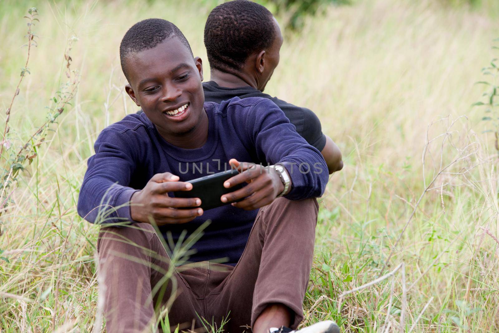 Close up of two young men leaning against each other and sitting on the grass in the park. one seen from behind and the other seen from the front uses a mobile phone. Concept of friendship men.