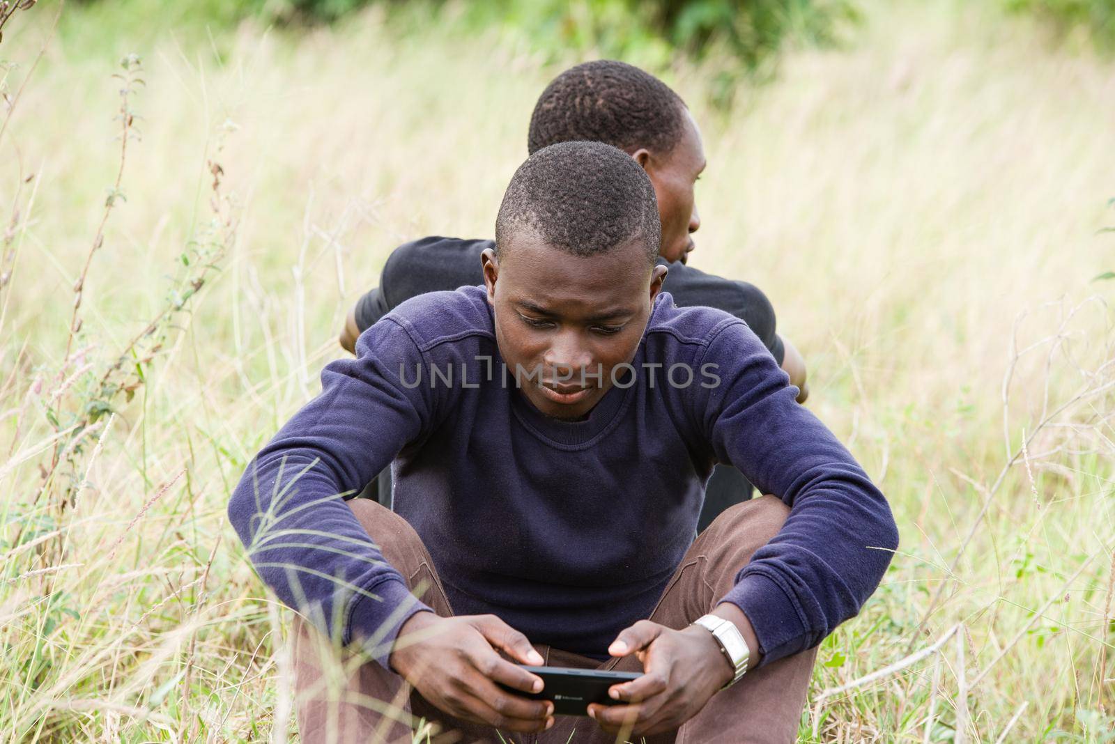 Close up of two young men leaning against each other and sitting on the grass in the park. one seen from behind and the other seen from the front uses a mobile phone. Concept of friendship men.