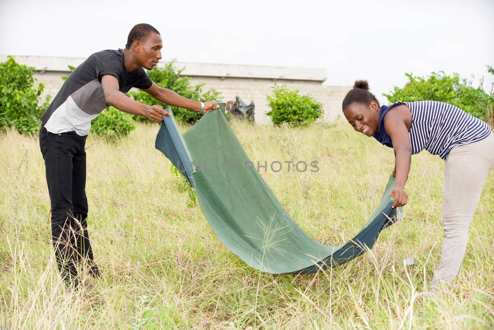 young african couple stretching blanket in a park smiling.