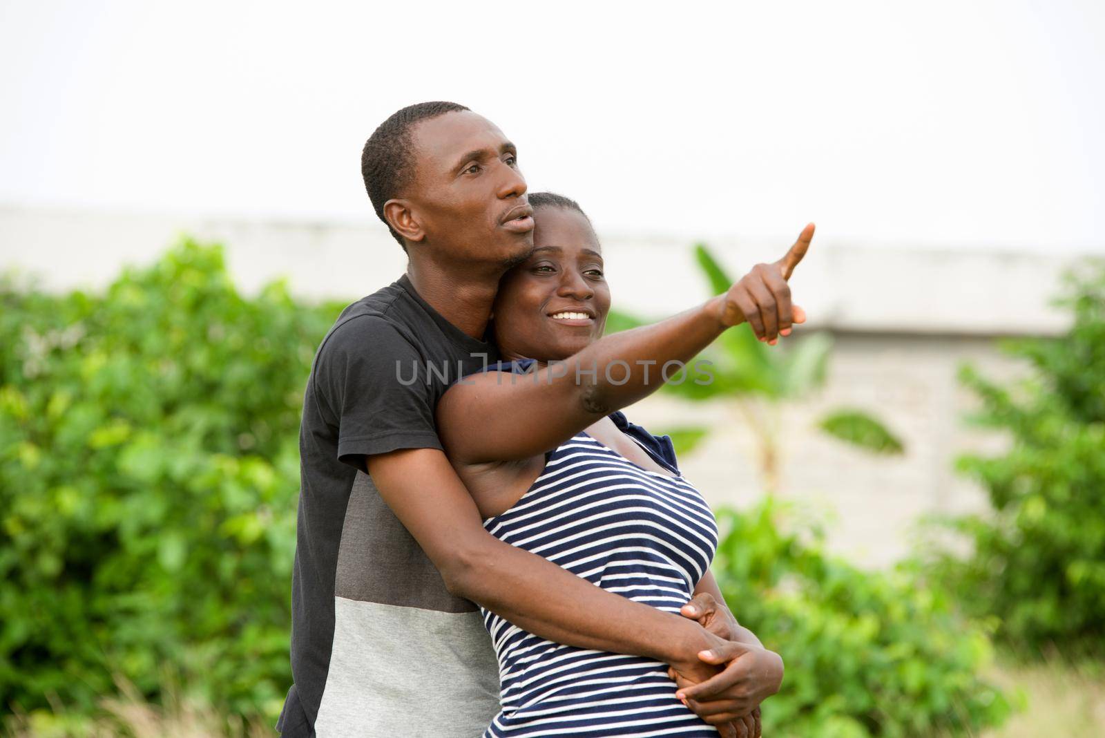 Young couple in a good mood hugging standing at the park in the middle of the day. The woman is pointing her finger elsewhere.