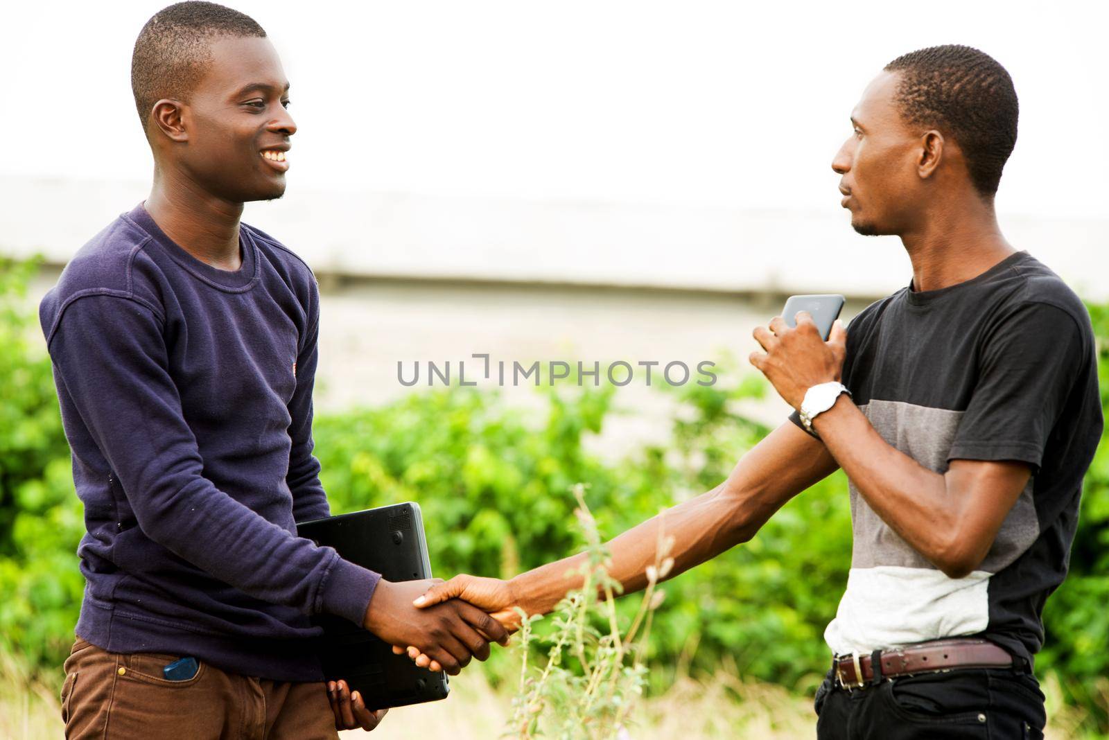 happy meeting and greeting between two young student standing in a field during the day and holding a computer in the hands