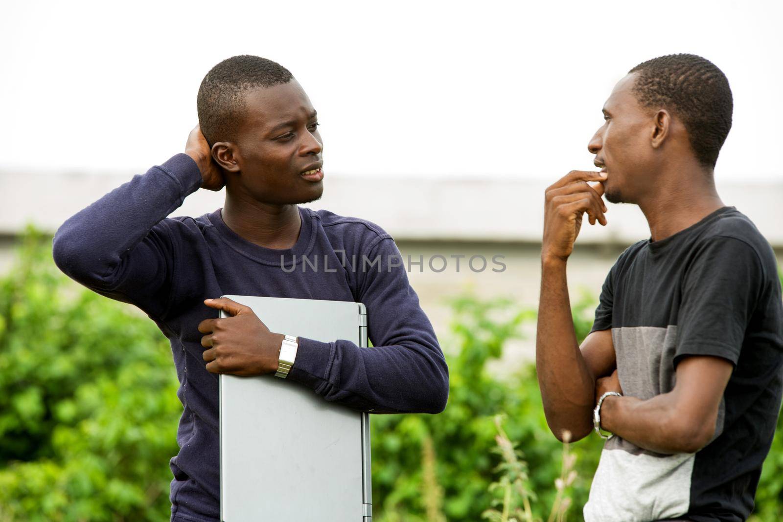 two young student chatting and laughing standing outdoors with a computer in their hands